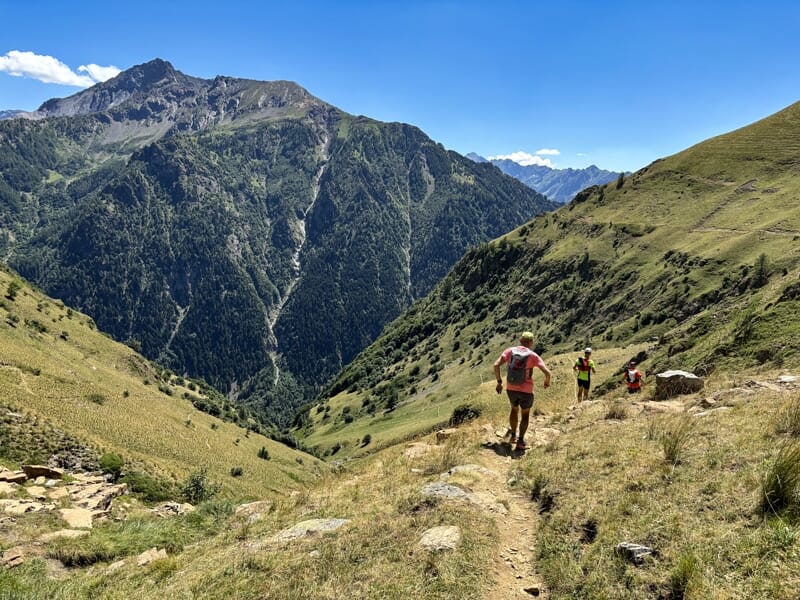 Des coureurs descendent un sentier étroit en montagne, entourés de collines verdoyantes et de forêts.