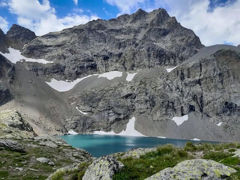 Un lac de montagne turquoise entouré de rochers et de sommets enneigés sous un ciel partiellement nuageux.