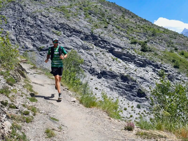 Un coureur en tenue verte court sur un chemin de montagne rocheux avec des falaises en arrière-plan.