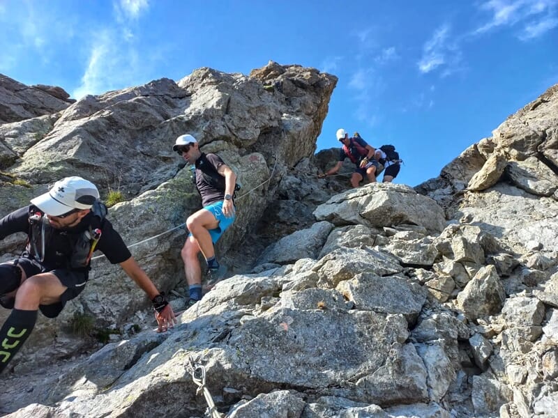 Des coureurs escaladent une pente rocheuse escarpée dans un paysage de montagne sous un ciel bleu.