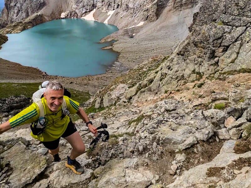 Un homme souriant grimpe une pente rocheuse avec un lac turquoise en contrebas et des montagnes en arrière-plan.