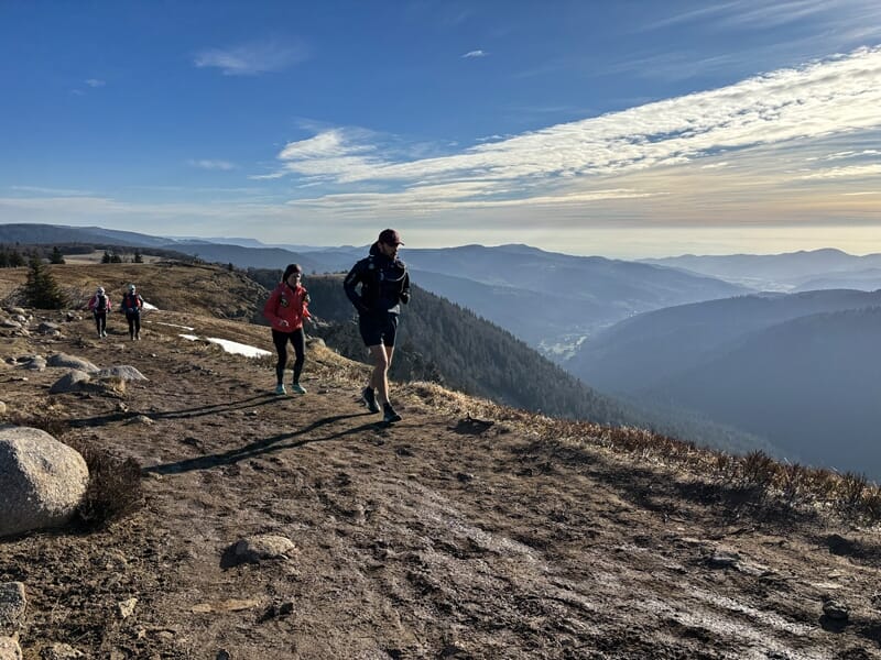 Des coureurs en trail sur un sentier de montagne au Col de la Schlucht sous un ciel bleu clair et lumineux.