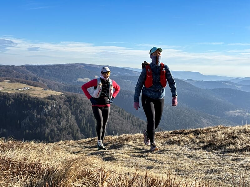 Deux coureurs en tenue de trail marchant sur un chemin de montagne avec une vue panoramique sur les vallées.