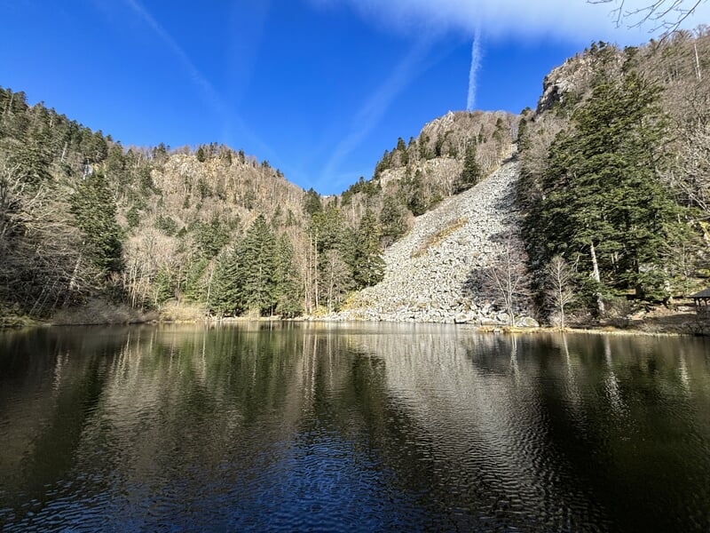 Petit lac entouré d'arbres et d'une pente rocheuse sous un ciel bleu avec quelques nuages blancs.