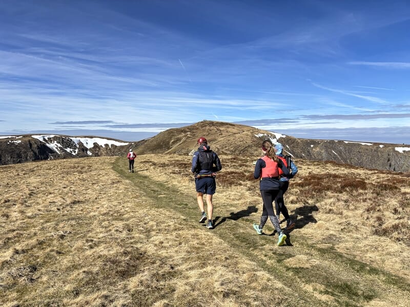 Trois coureurs en trail progressant sur un sentier herbeux en montagne avec un ciel bleu et nuages.