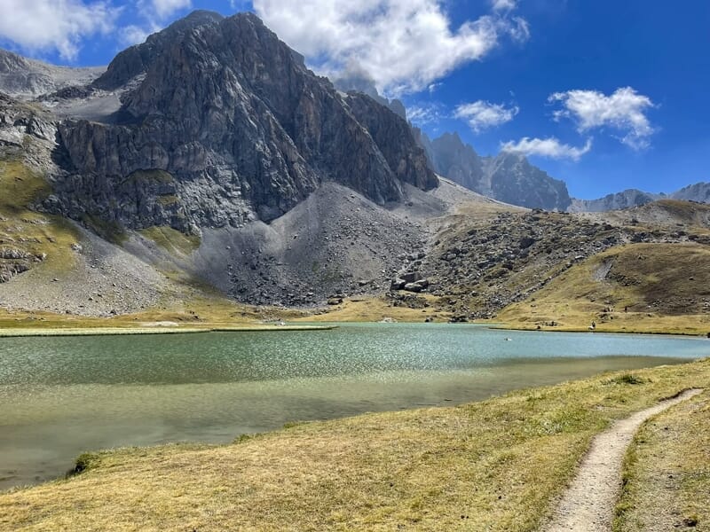 Lac de montagne entouré de rochers et d'herbe sèche sous un ciel bleu avec quelques nuages blancs.