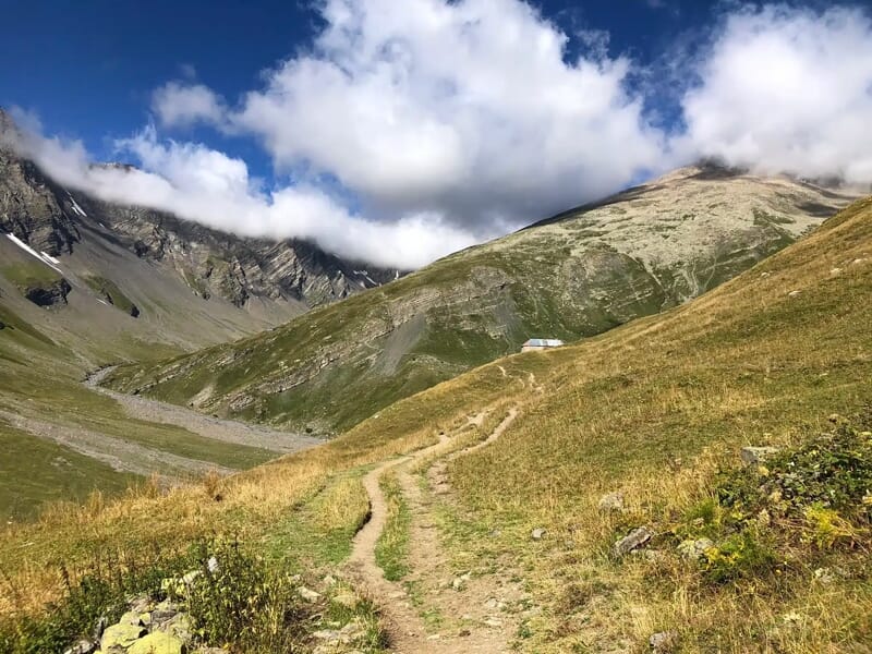 Sentier de randonnée en montagne avec herbe jaune et verte, sous un ciel partiellement nuageux.