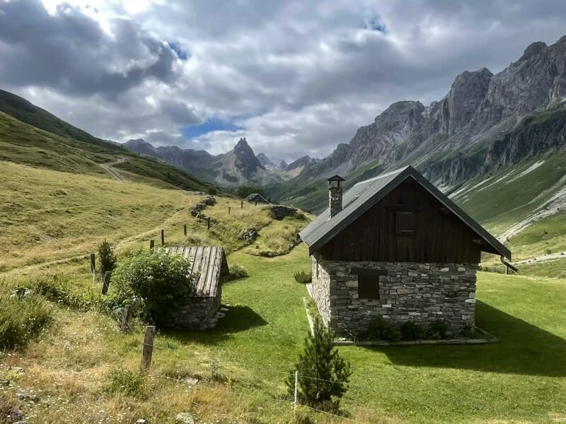 Chalet en pierre dans une vallée verte entourée de montagnes sous un ciel nuageux.