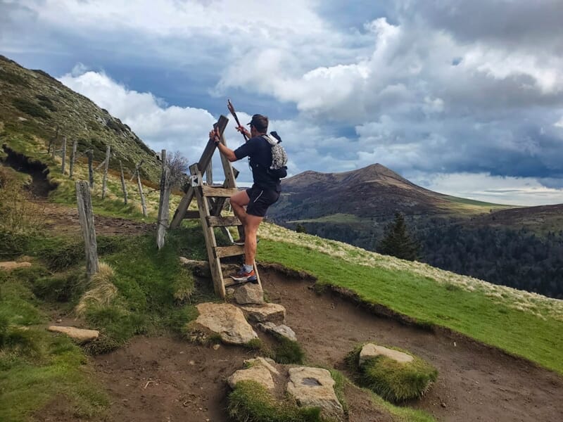 Un coureur franchit une barrière en bois sur un sentier de montagne avec un paysage vallonné en arrière-plan.