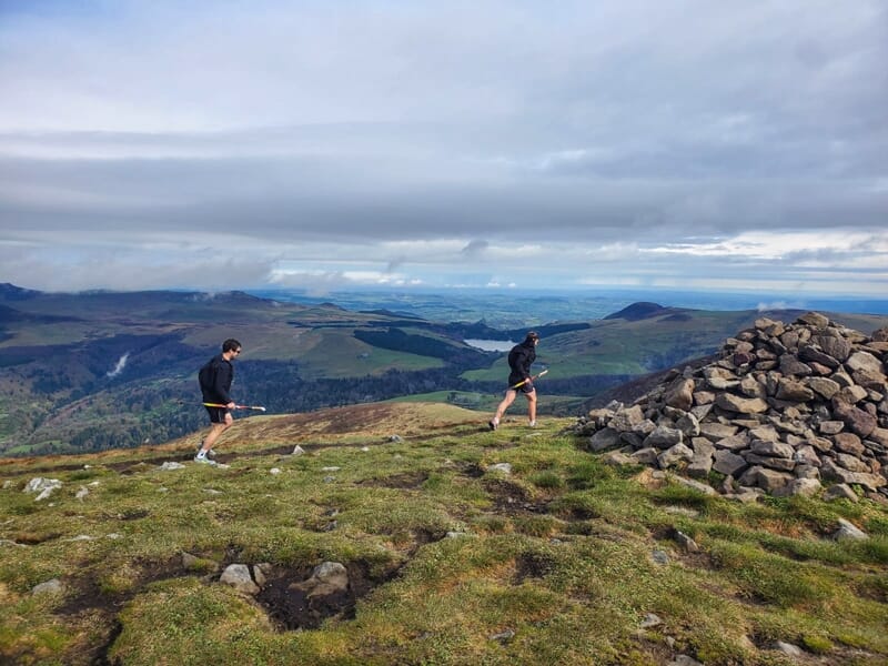 Deux coureurs sur un sommet herbeux près d'un cairn de pierres sous un ciel nuageux.