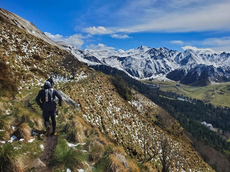 Un coureur descend un sentier étroit en montagne avec des sommets enneigés en arrière-plan.