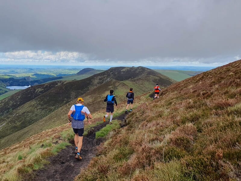 Quatre coureurs en tenue sportive descendent un sentier de montagne sous un ciel nuageux.