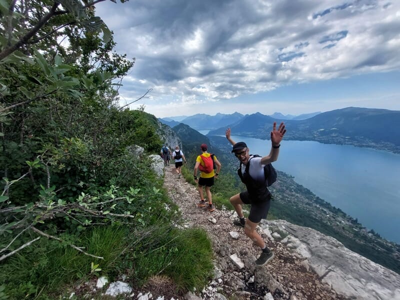 Groupe de coureurs sur un sentier étroit en montagne avec vue sur un lac et des collines verdoyantes.