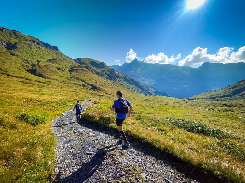 Deux coureurs sur un chemin de montagne entouré d'herbe verte sous un ciel bleu avec quelques nuages.