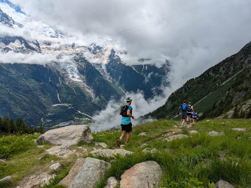 Coureurs descendant un sentier en montagne avec des nuages bas et des sommets rocheux en arrière-plan.