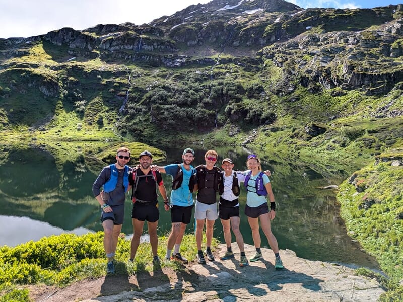 Groupe de six personnes posant souriantes devant un lac de montagne entouré de végétation luxuriante.
