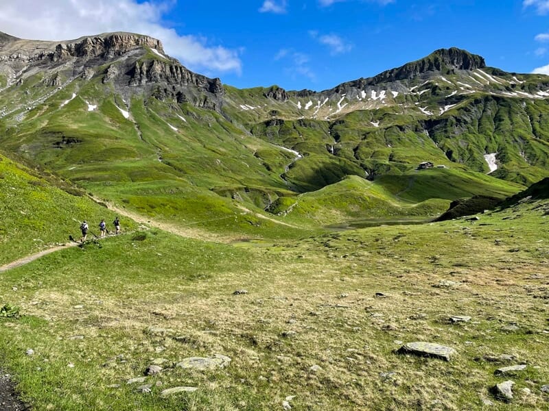 Coureurs sur un large sentier de montagne avec collines verdoyantes et ciel bleu lumineux en arrière-plan.