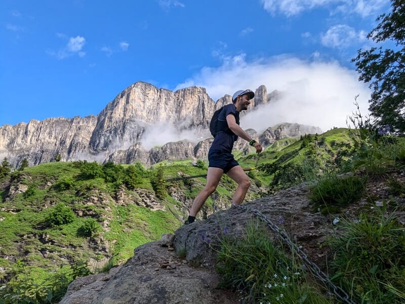 Homme courant sur un sentier rocheux en montagne avec un massif montagneux partiellement couvert de nuages.