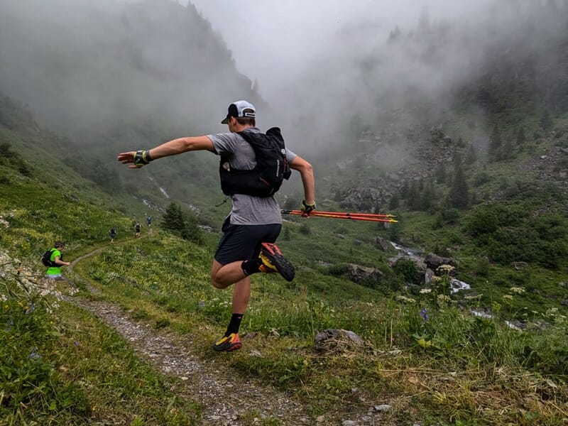 Coureur sautant sur un sentier de montagne entouré de végétation dense et de nuages bas dans un paysage vert.