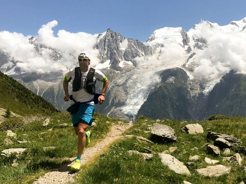 Un coureur en tenue sportive court sur un sentier de montagne avec un paysage alpin enneigé en arrière-plan.