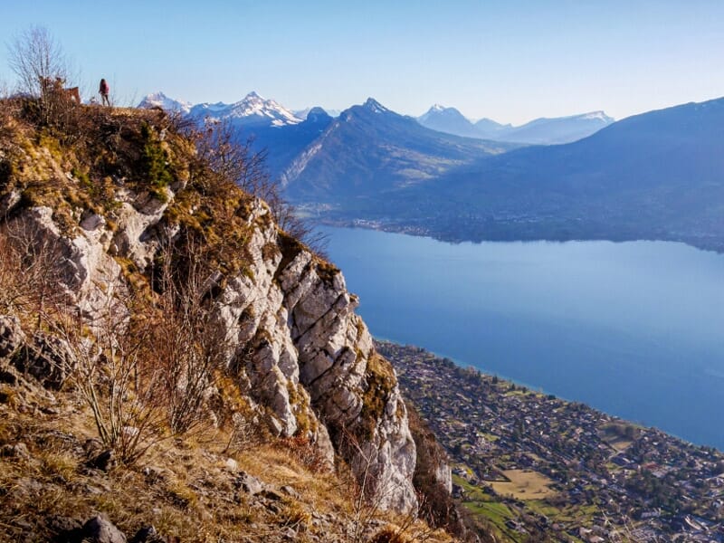 Vue panoramique sur un lac entouré de montagnes rocheuses et de végétation, sous un ciel clair et lumineux.