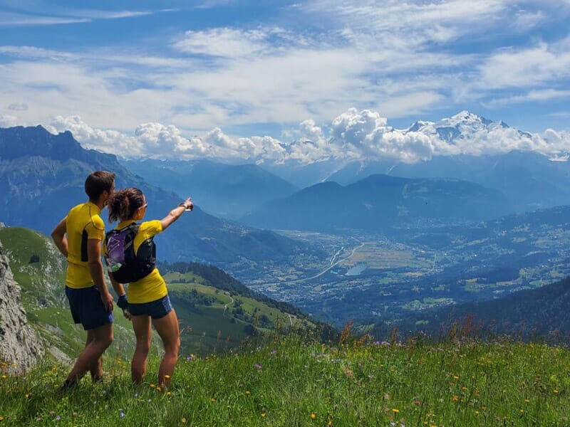 Deux randonneurs en tenue jaune admirent la vue sur une vallée montagneuse sous un ciel partiellement nuageux.