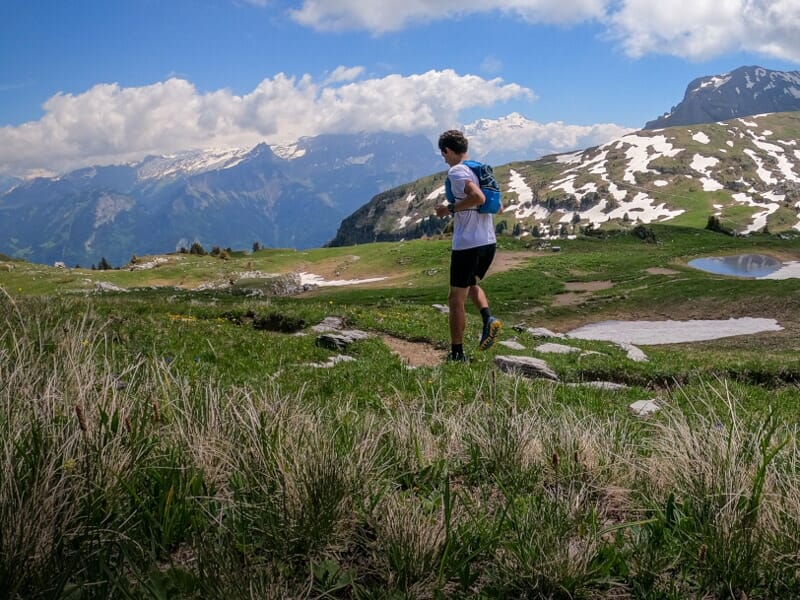 Un homme court sur un sentier de montagne avec des plaques de neige et un ciel bleu parsemé de nuages.