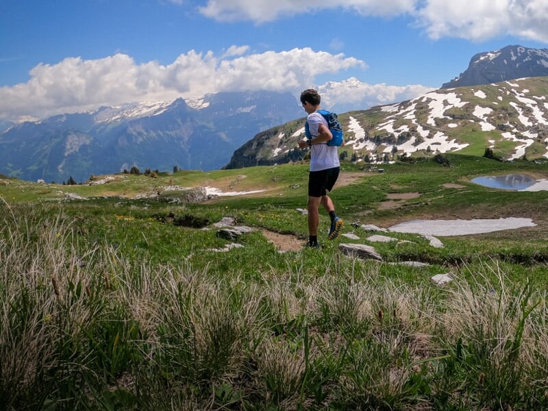 Un homme court sur un sentier de montagne avec des plaques de neige et un ciel bleu parsemé de nuages.