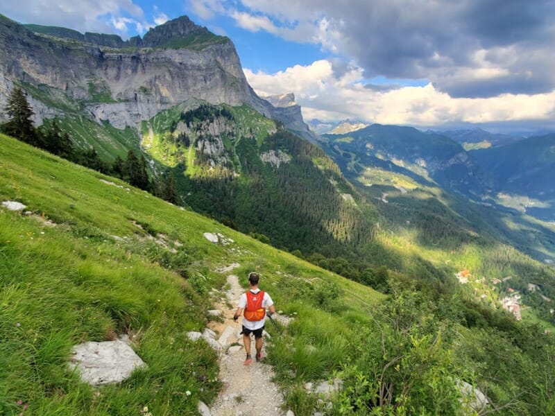 Un coureur descend un sentier étroit en montagne, entouré de prairies vertes et de falaises rocheuses.