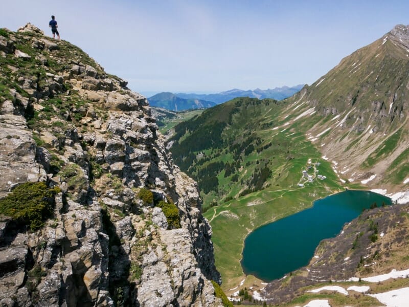 Un randonneur se tient au sommet d'une falaise rocheuse dominant un lac bleu entouré de montagnes verdoyantes.