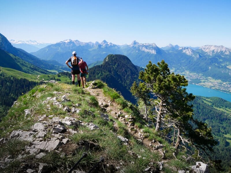 Deux personnes marchent sur un sentier étroit en crête avec une vue dégagée sur les montagnes et un lac au loin.