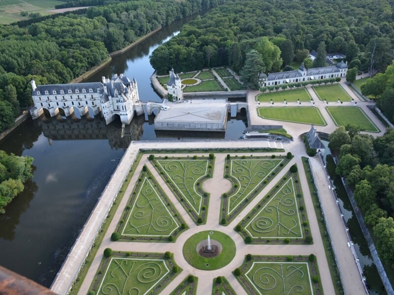 Vue aérienne du domaine comprenant un château et ses jardins à la française, avec un canal et des bois environnants, baigné dans la lumière du jour