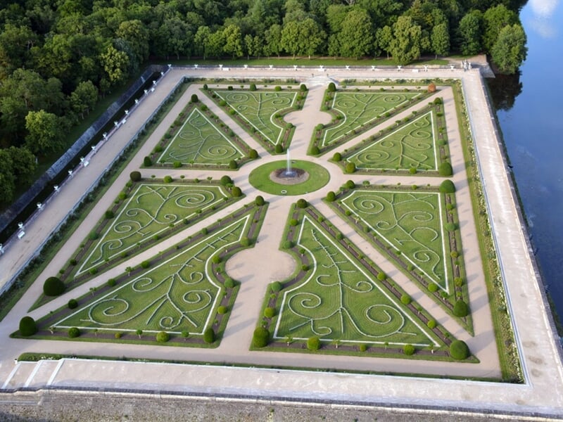 Jardin à la française avec des parterres géométriques symétriques, arbustes taillés et allées gravillonnées, bordé d'arbres et d'un canal