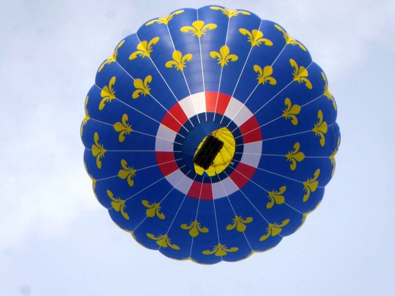 Montgolfière bleu avec des fleurs de lys jaunes et un motif de cocarde tricolore au centre, vue de dessous contre un ciel clair
