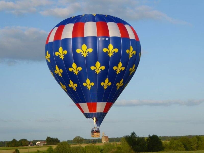 Montgolfière bleu ornée de fleurs de lys jaunes et de bandes tricolores, flottant dans le ciel clair au-dessus d'un paysage rural