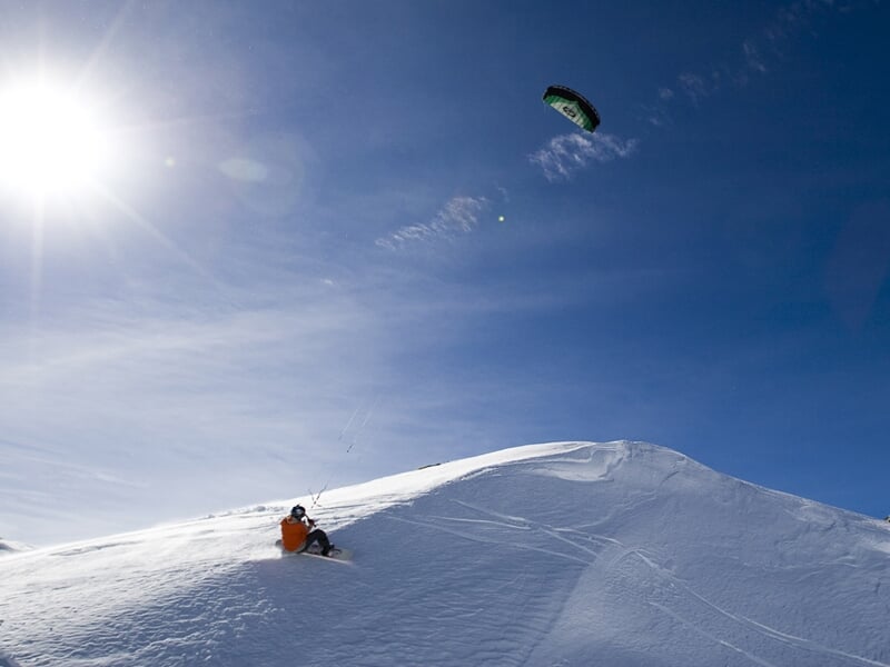 Personne pratiquant le snowkite sur une colline enneigée sous un ciel bleu avec un soleil éclatant.