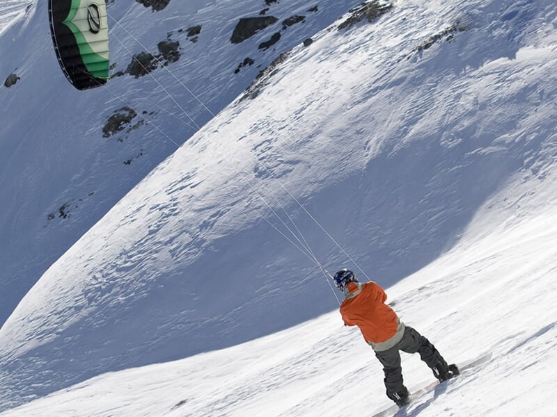 Homme en tenue d'hiver pilotant une aile de snowkite sur une pente enneigée en montagne.