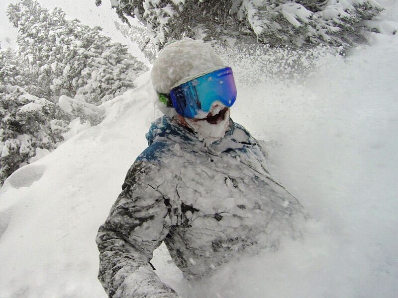 Skieur couvert de neige poudreuse avec lunettes de protection dans un environnement forestier enneigé.