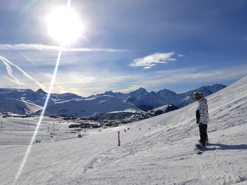 Skieur debout sur une piste enneigée avec des montagnes en arrière-plan sous un ciel ensoleillé.