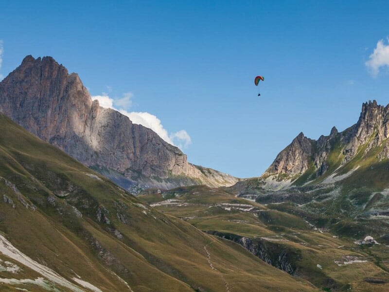 Un parapentiste vole au-dessus des montagnes rocheuses et vallonnées sous un ciel bleu clair avec quelques nuages.