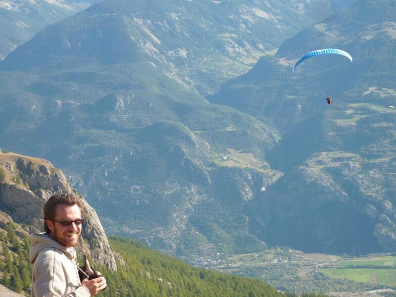 Un homme souriant avec des lunettes observe un parapente rouge en vol dans une vallée montagneuse verdoyante.