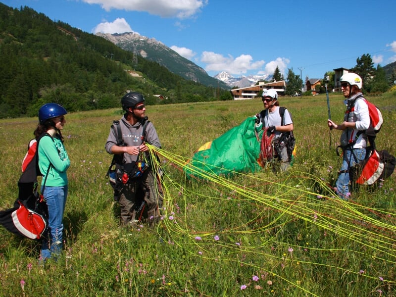 Quatre personnes en tenue de parapente discutent dans un champ vert avec des montagnes en arrière-plan.