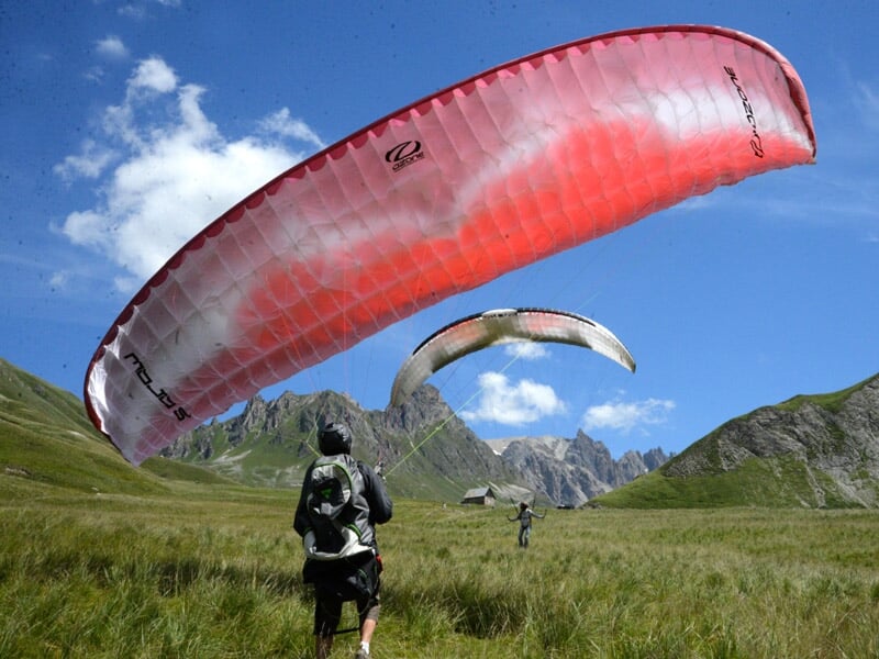 Un parapentiste prépare son aile rouge et blanche dans un champ herbeux sous un ciel bleu avec des montagnes.