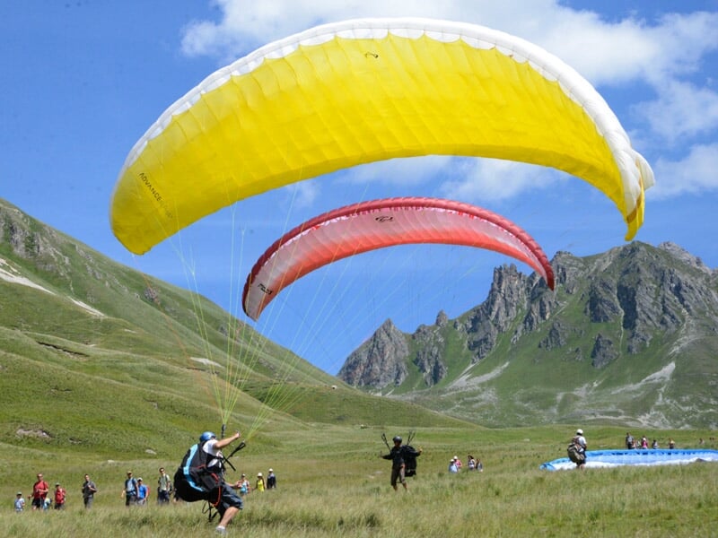 Deux parapentes jaunes et rouges s'élèvent dans un champ vert avec plusieurs personnes et montagnes en arrière-plan.