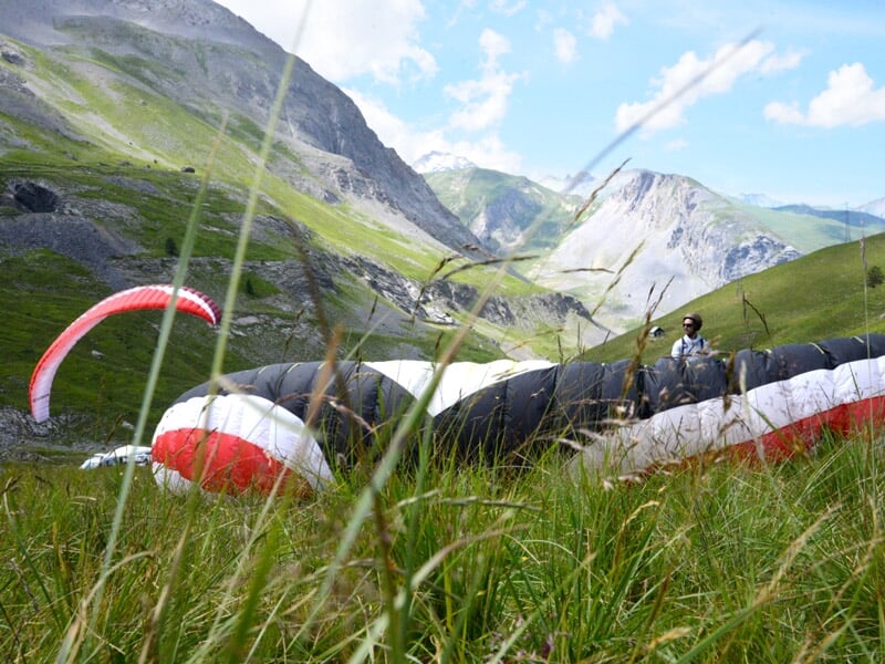 Plusieurs parapentistes avec des ailes colorées s'élancent dans un champ de montagne verdoyant sous un ciel bleu.