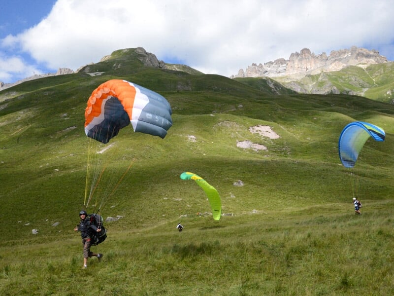 Un parapente noir et rouge est posé dans l'herbe avec des montagnes en arrière-plan sous un ciel clair.
