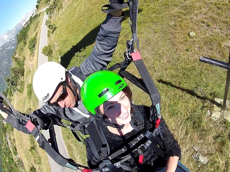 Un homme et une femme en tandem parapente sourient en vol, la femme porte un casque vert et le ciel est dégagé.