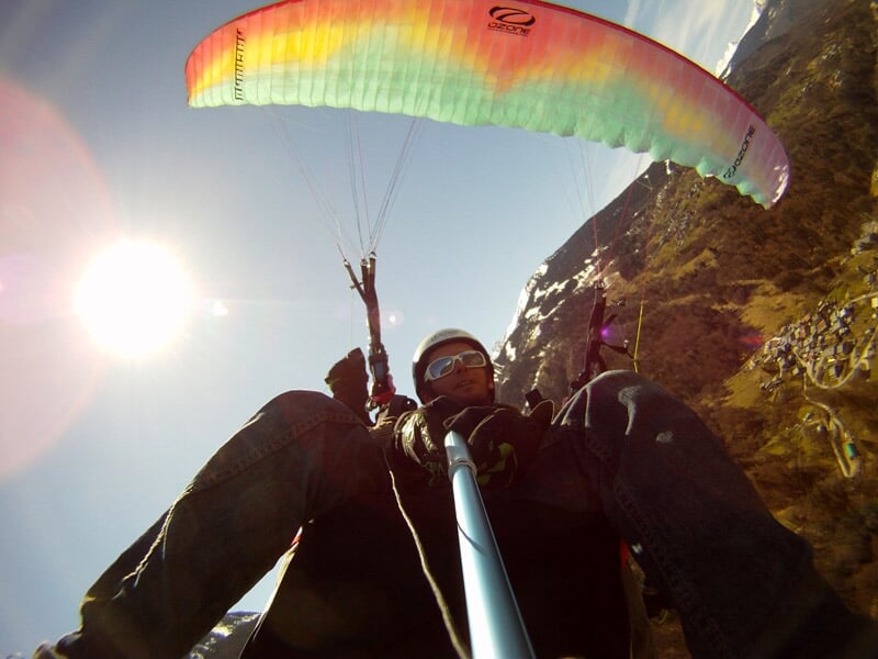Un homme en parapente biplace prend un selfie en vol avec le soleil brillant et des montagnes en arrière-plan.