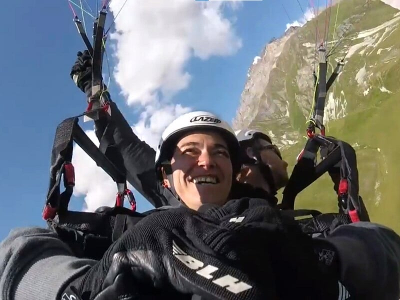 Deux personnes en parapente biplace souriantes volent au-dessus d'une vallée verte avec des montagnes sous un ciel bleu.