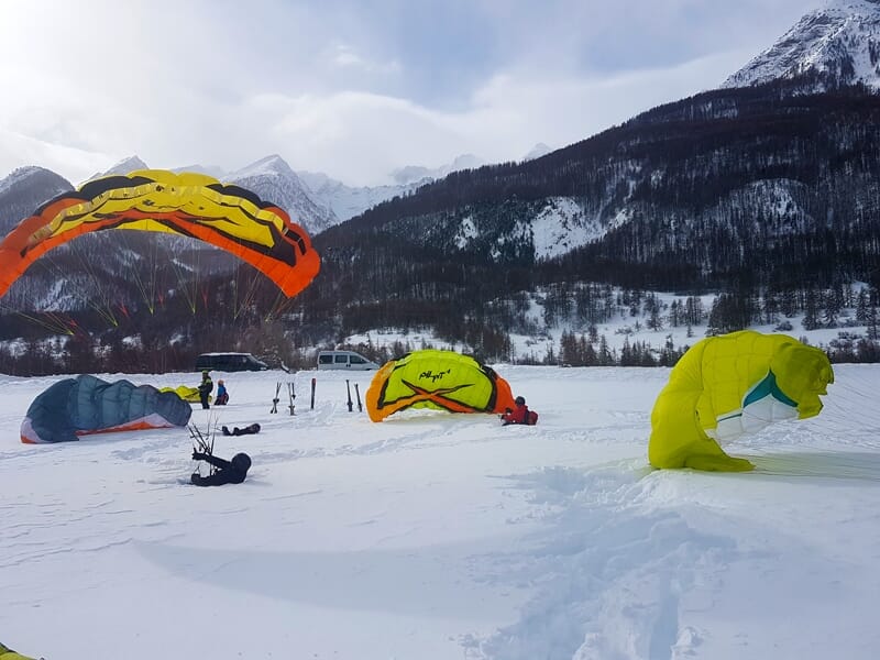 Personnes préparant leur parapente sur la neige dans une vallée entourée de montagnes enneigées sous un ciel nuageux.